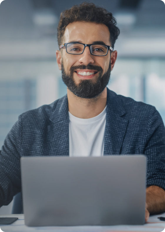 Homem de óculos e barba, sorridente, sentado em uma mesa com um laptop.