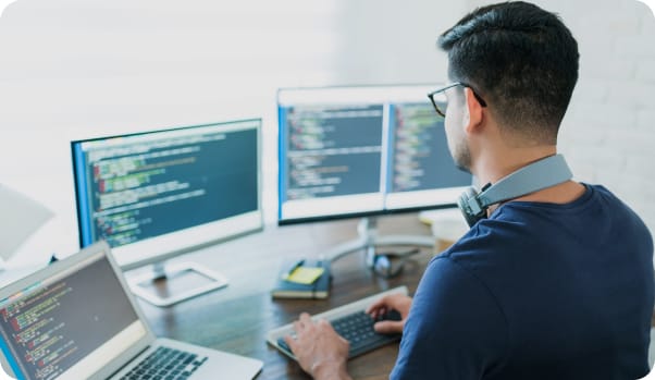 Homem trabalhando em três computadores, focado em atividades relacionadas a soluções web.